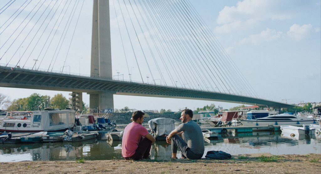 Still from "The Lawyer" - Marius & Ali sit together alongside the river, with a suspension bridge in the background